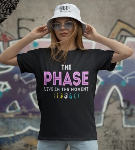 Güneş şapka ve renkli tişört ile genç kadın, ay fazları temalı 'The Phase' yazılı tişörtü giyiyor, şehir grafiti arka planıyla yoga ve doğa temalı modern kıyafet tarzı sergiliyor.